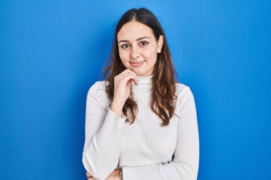 Mavi arka planda, elleri çenesinde soru soran, düşünceli bir İspanyol kadın. Gülümseyen ve düşünceli bir yüz. şüphe kavramı. 
