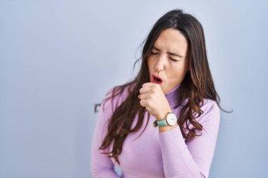 Mavi arka planda duran esmer kadın kendini kötü hissediyor ve soğuk algınlığı veya bronşitin belirtisi olarak öksürüyor. sağlık hizmeti kavramı. 