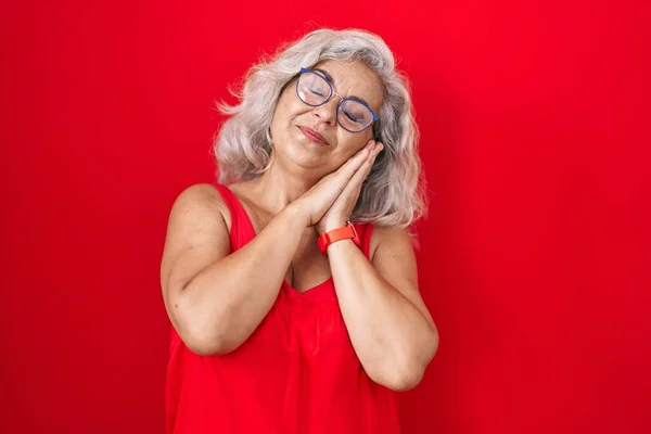 Middle age woman with grey hair standing over red background sleeping tired dreaming and posing with hands together while smiling with closed eyes.