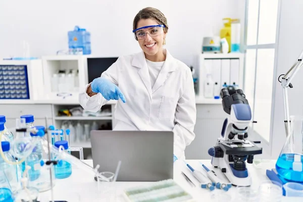 Young hispanic woman wearing scientist uniform working at laboratory smiling happy pointing with hand and finger