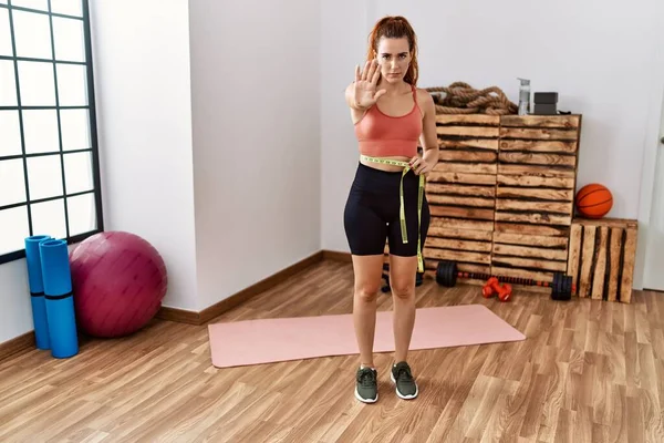 Young redhead woman using tape measure measuring waist at the gym with open hand doing stop sign with serious and confident expression, defense gesture