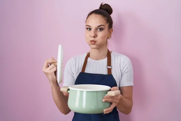 Young Hispanic Girl Wearing Apron Holding Cooking Pot Puffing Cheeks — Zdjęcie stockowe