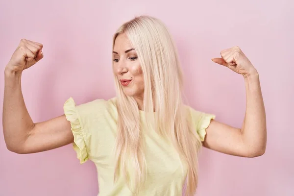 Caucasian woman standing over pink background showing arms muscles smiling proud. fitness concept.