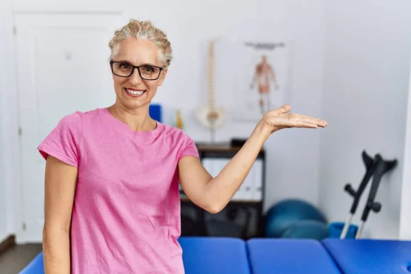 Middle age blonde woman at pain recovery clinic smiling cheerful presenting and pointing with palm of hand looking at the camera.
