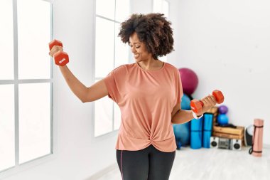 Genç Afrikalı Amerikalı kadın gülümsüyor spor merkezinde halterleri kullanarak kendine güven eğitimi alıyor