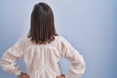 Orta yaşlı İspanyol bir kadın mavi arka planda dikiliyor geriye bakıyor kolları vücudunda. 