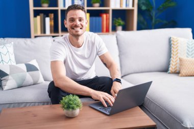Genç İspanyol adam evdeki koltukta dizüstü bilgisayar kullanıyor.