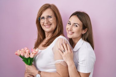 Hispanic mother and daughter holding pink tulips bouquet looking positive and happy standing and smiling with a confident smile showing teeth 
