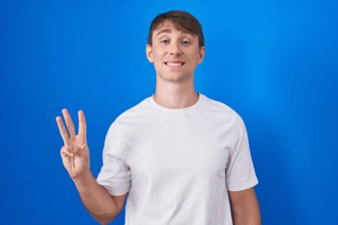 Caucasian blond man standing over blue background showing and pointing up with fingers number three while smiling confident and happy. 