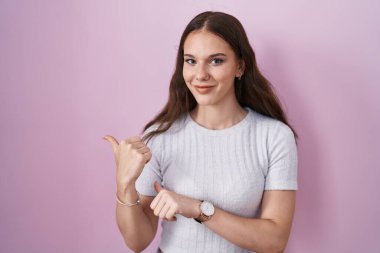 Young hispanic girl standing over pink background pointing to the back behind with hand and thumbs up, smiling confident 