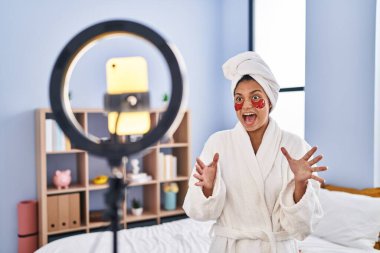 Koyu renk saçlı İspanyol bir kadın akıllı telefonuyla güzellik dersi kaydediyor zaferi mutlu bir gülümsemeyle ve ellerini kaldırarak kutluyor. 