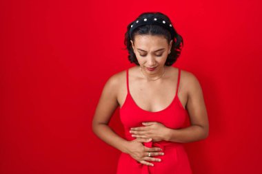 Young hispanic woman standing over red background with hand on stomach because indigestion, painful illness feeling unwell. ache concept. 
