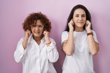 Hispanic mother and daughter wearing casual white t shirt over pink background covering ears with fingers with annoyed expression for the noise of loud music. deaf concept. 