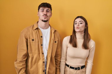 Young hispanic couple standing over yellow background looking at the camera blowing a kiss on air being lovely and sexy. love expression. 