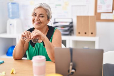 Orta yaşlı, gri saçlı, iş kadını. Ofisteki masada kendinden emin bir şekilde oturuyor.