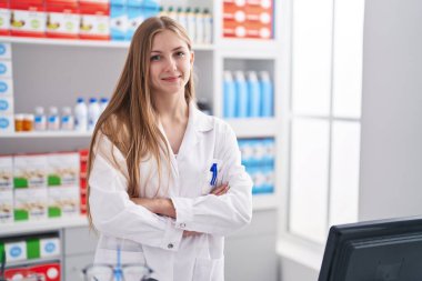 Young caucasian woman pharmacist smiling confident standing with arms crossed gesture at pharmacy