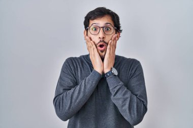 Handsome latin man standing over isolated background afraid and shocked, surprise and amazed expression with hands on face 