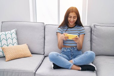 Young hispanic girl playing video game by smartphone at home