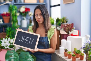 Hispanic young woman working at florist with open sign relaxed with serious expression on face. simple and natural looking at the camera. 