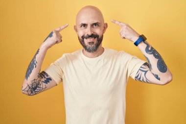 Sarı arka planda dikilen İspanyol dövmeli adam iki eliyle başını gösteren gülümseme, harika bir fikir ya da düşünce, iyi bir hafıza 