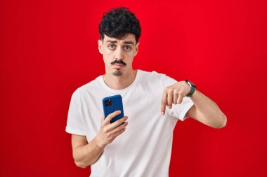 Hispanic man using smartphone over red background pointing down looking sad and upset, indicating direction with fingers, unhappy and depressed. 