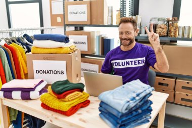 Middle age man wearing volunteer t shirt working with laptop showing and pointing up with fingers number two while smiling confident and happy. 