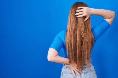 Redhead woman standing over blue background backwards thinking about doubt with hand on head 