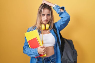 Young blonde woman wearing student backpack and holding books confuse and wondering about question. uncertain with doubt, thinking with hand on head. pensive concept. 