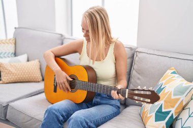 Genç sarışın kadın evdeki kanepede oturmuş klasik gitar çalıyor.