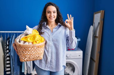 Young brunette woman holding laundry basket doing ok sign with fingers, smiling friendly gesturing excellent symbol 