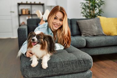 Beyaz bir kadın, evinde köpekle kanepede uzanmış kendinden emin bir şekilde gülümsüyor.