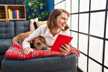 Kafkasyalı genç bir kadın evdeki köpekle birlikte kanepeye uzanmış dokunmatik ped kullanıyor.