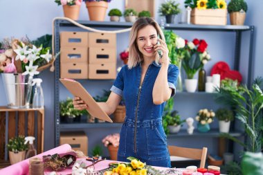 Young woman florist talking on smartphone reading clipboard at florist