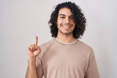 Beyaz arka planda duran kıvırcık saçlı İspanyol adam bir numarayı gösterip işaret ederken kendinden emin ve mutlu gülümsüyor.. 