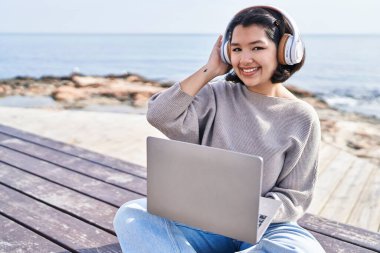 Young woman using laptop and headphones sitting on bench at seaside