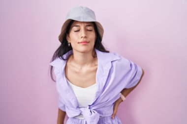 Young hispanic woman standing over pink background wearing hat suffering of backache, touching back with hand, muscular pain 