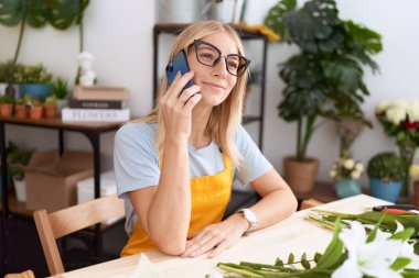 Young blonde woman florist smiling confident talking on smartphone at flower shop