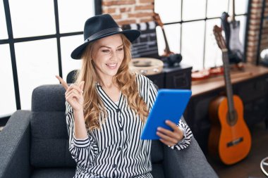 Beautiful blonde woman at music studio using tablet smiling happy pointing with hand and finger to the side 