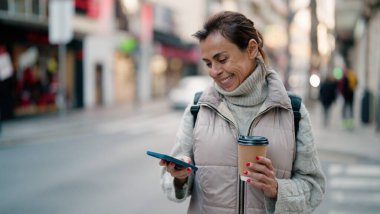 Orta yaşlı İspanyol kadın akıllı telefon kullanıyor. Sokakta kahve içiyor.