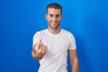 Young caucasian man standing over blue background beckoning come here gesture with hand inviting welcoming happy and smiling 