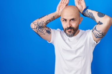 Mavi arka planda dikilen İspanyol dövmeli adam avuçlarında alaycı ve şüpheci bakışlarla tavşan kulağı hareketi yapıyor. Paskalya tavşanı konsepti. 