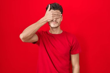 Young hispanic man standing over red background covering eyes with hand, looking serious and sad. sightless, hiding and rejection concept 
