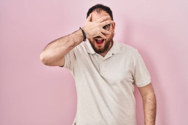 Plus size hispanic man with beard standing over pink background peeking in shock covering face and eyes with hand, looking through fingers with embarrassed expression. 