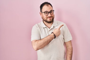 Plus size hispanic man with beard standing over pink background cheerful with a smile on face pointing with hand and finger up to the side with happy and natural expression 
