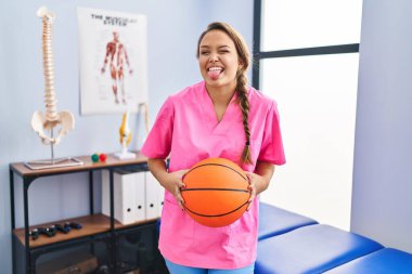 Young hispanic woman working at physiotherapy clinic holding basketball ball sticking tongue out happy with funny expression. 