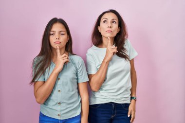 Young mother and daughter standing over pink background thinking concentrated about doubt with finger on chin and looking up wondering 