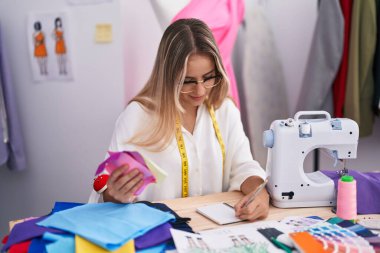 Young blonde woman tailor writing on notebook holding cloth at clothing shop