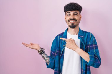 Young hispanic man with beard standing over pink background amazed and smiling to the camera while presenting with hand and pointing with finger. 