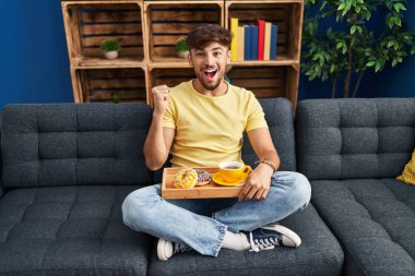 Arab man with beard sitting on the sofa at home eating breakfast screaming proud, celebrating victory and success very excited with raised arms 