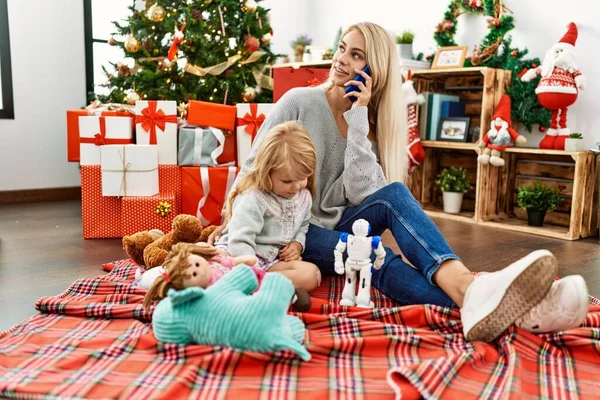 Mother and daughter playing with toys and talking on the smartphone sitting by christmas tree at home